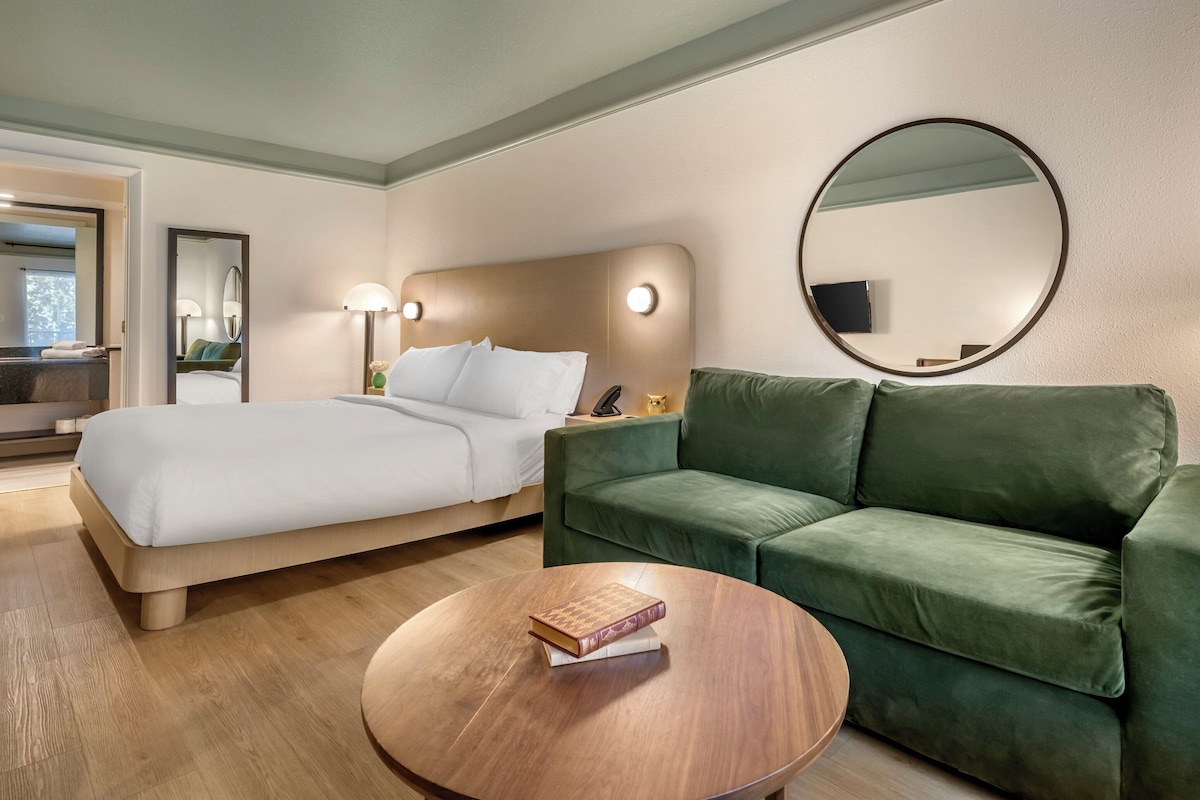Camera d'albergo con un letto, un divano verde, un tavolino rotondo con libri, uno specchio tondo e una TV a schermo piatto montata a parete.