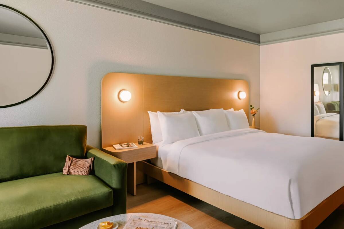 Una habitación de hotel con una cama grande, un sillón verde, una mesita de noche con lámpara y un espejo redondo en la pared.