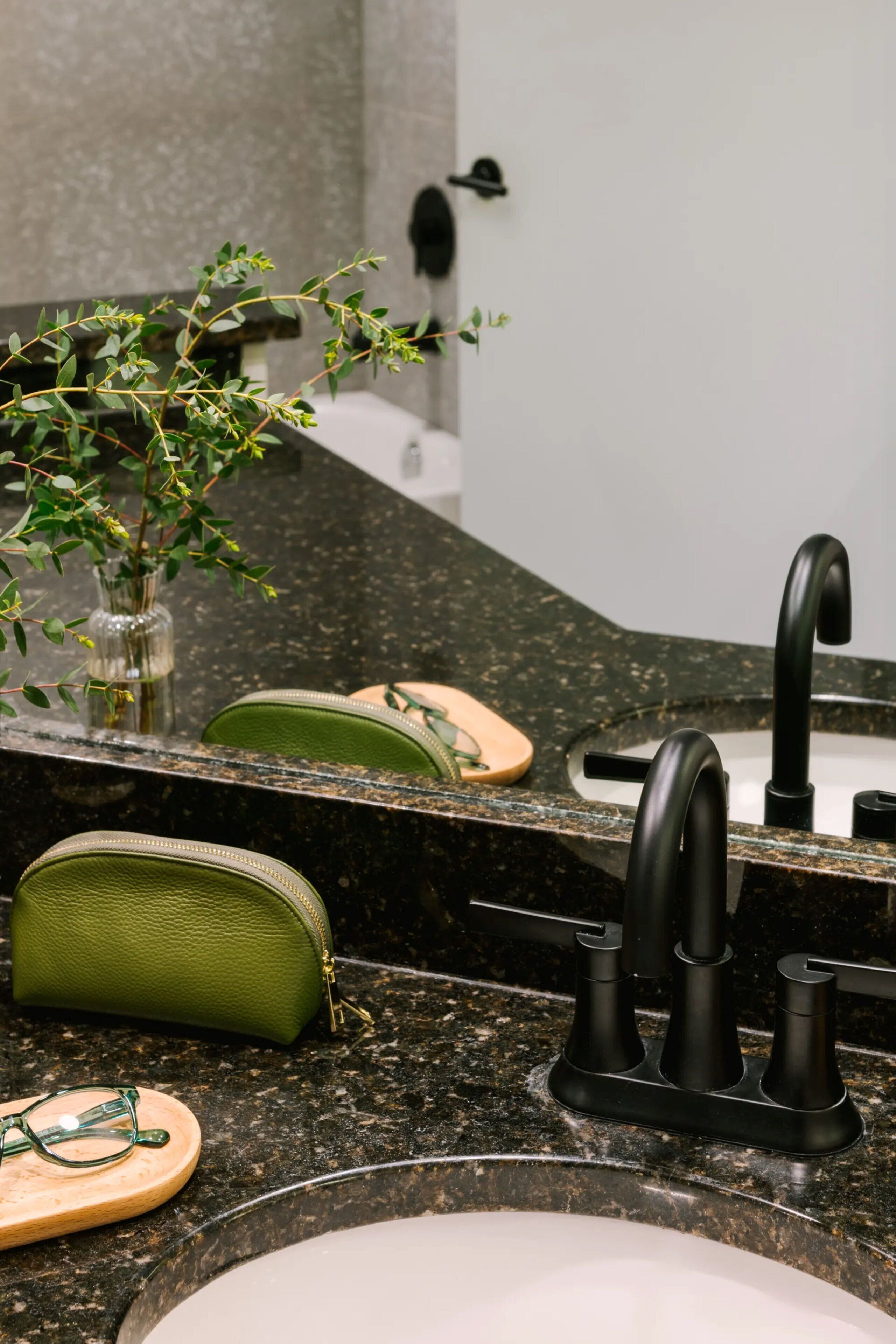 Un baño con encimera de granito, grifo negro, un bolso verde y una planta pequeña en un florero de vidrio.