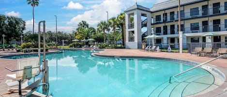 A swimming pool with a clear blue water, surrounded by lounge chairs and umbrellas, with a building in the background.