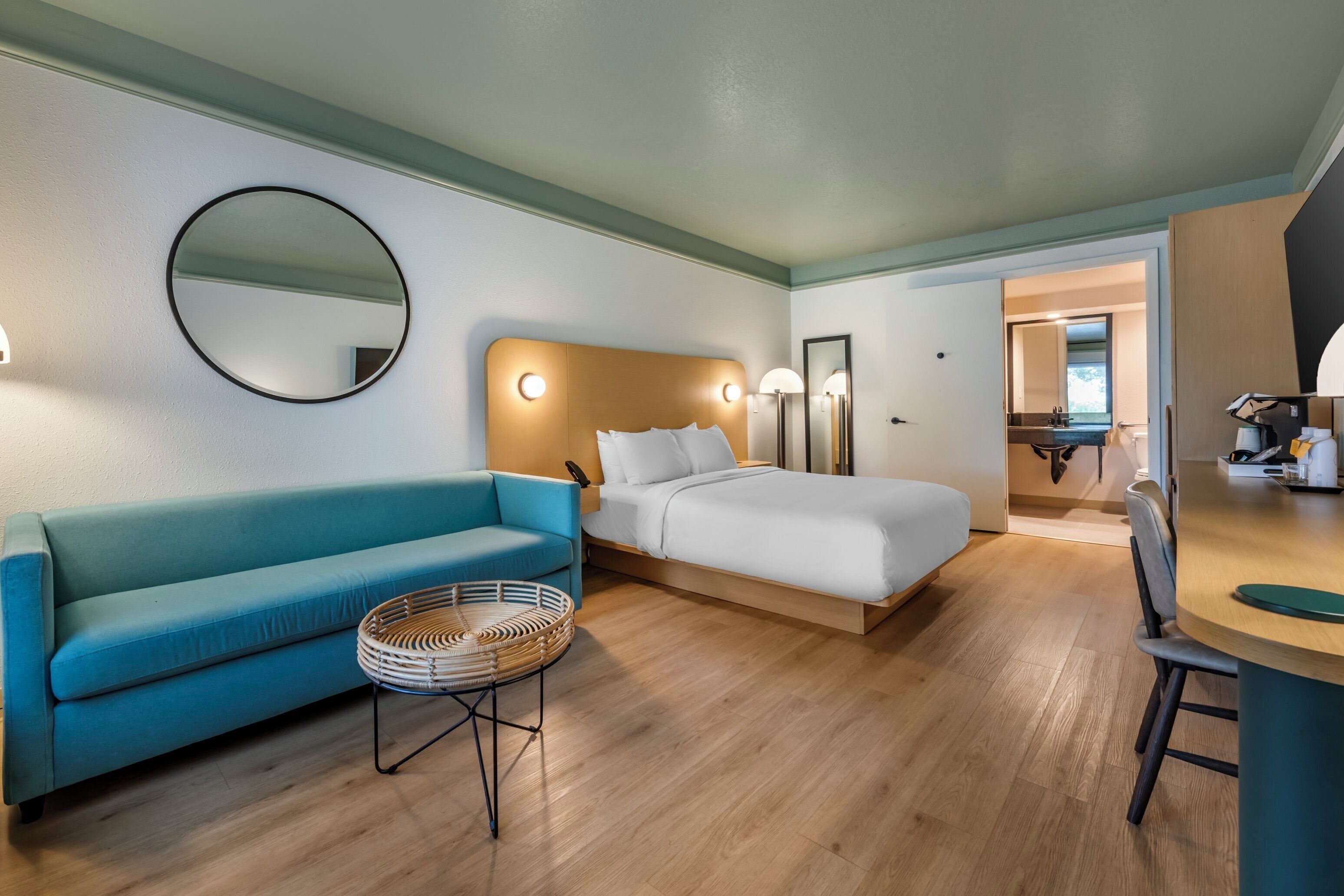 A modern hotel room with a bed, a blue sofa, a round wicker table, and a wooden dining table.