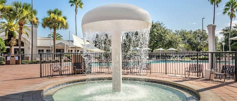 Una fuente con chorros de agua, rodeada por una piscina y sillones de descanso, con palmeras y un edificio al fondo.