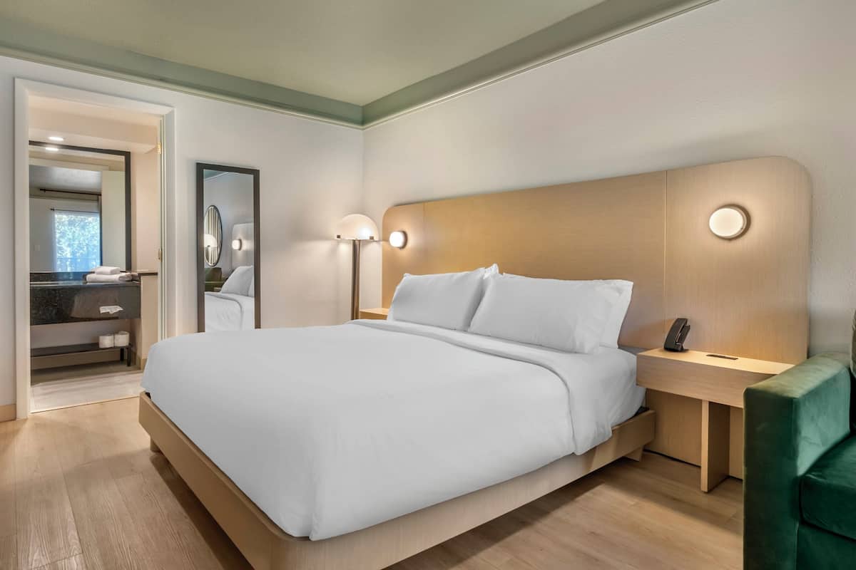 Habitació d'hotel moderna amb un llit gran, taules de nit, un mirall i una butaca verda.
