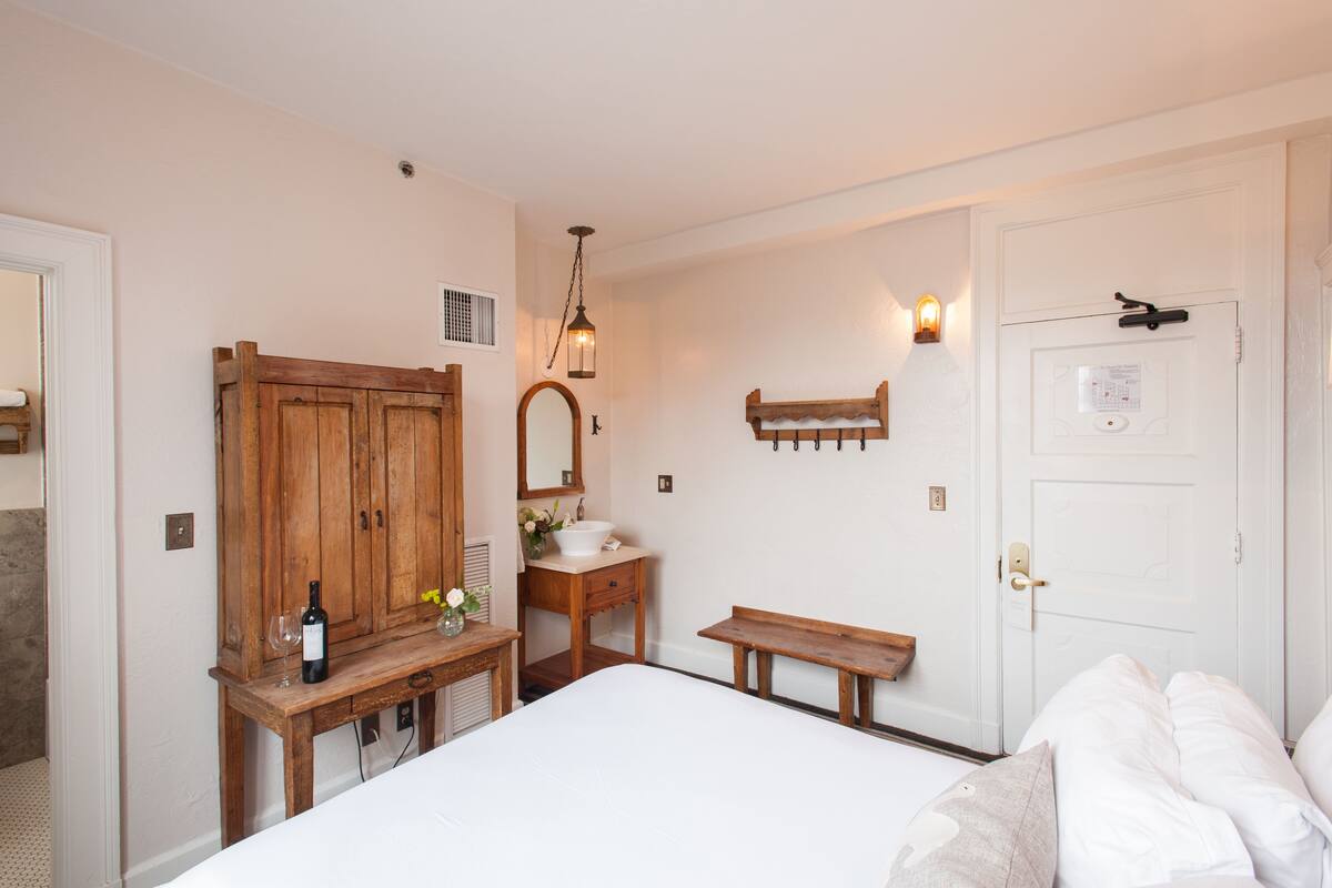 Un dormitorio con un armario de madera, una mesita de noche con una botella de vino y una copa, un banco y una puerta blanca con cerradura.