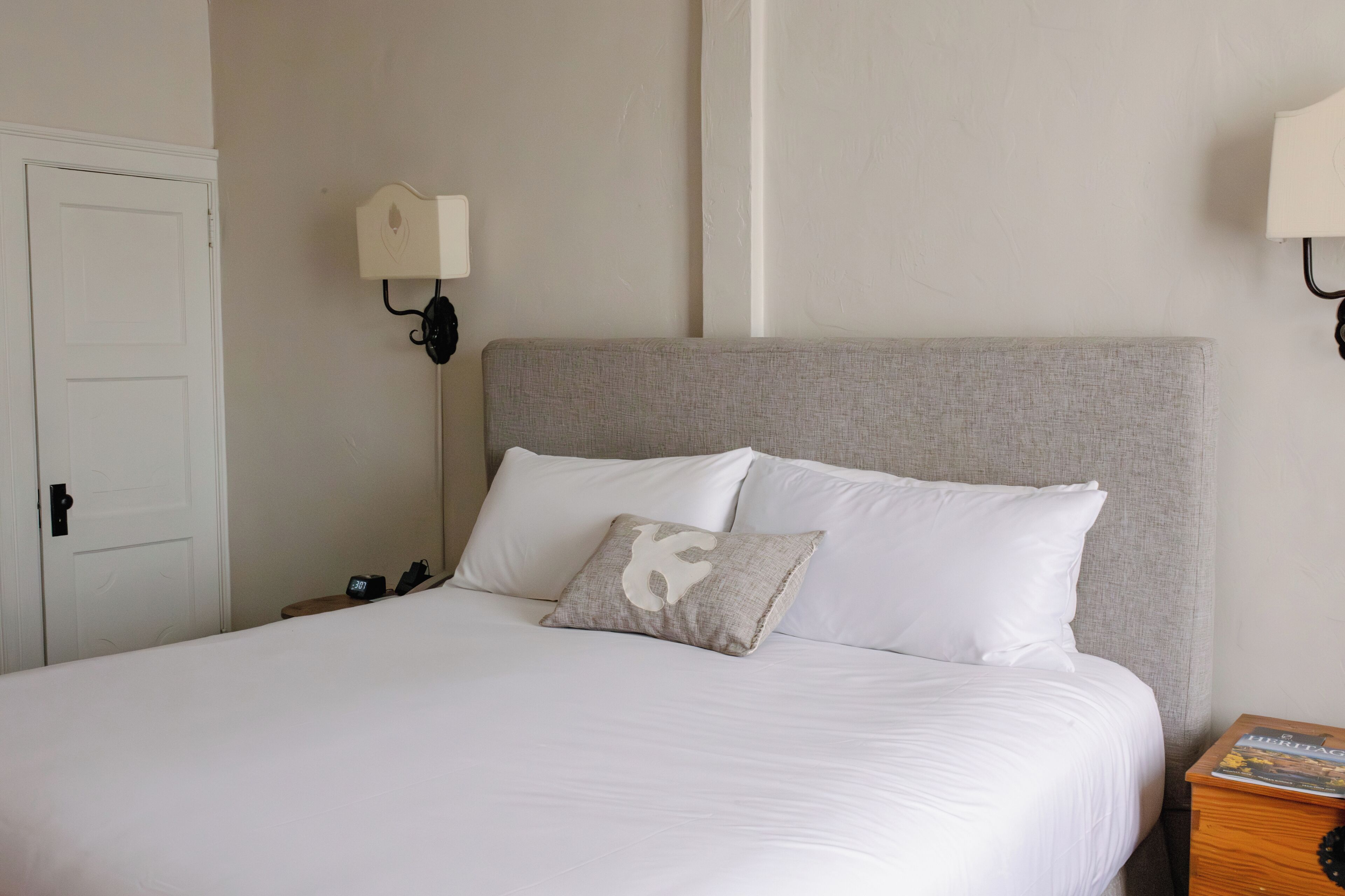 A neatly made bed with white bedding and a single decorative pillow, a bedside table with a book, and a wall-mounted lamp.