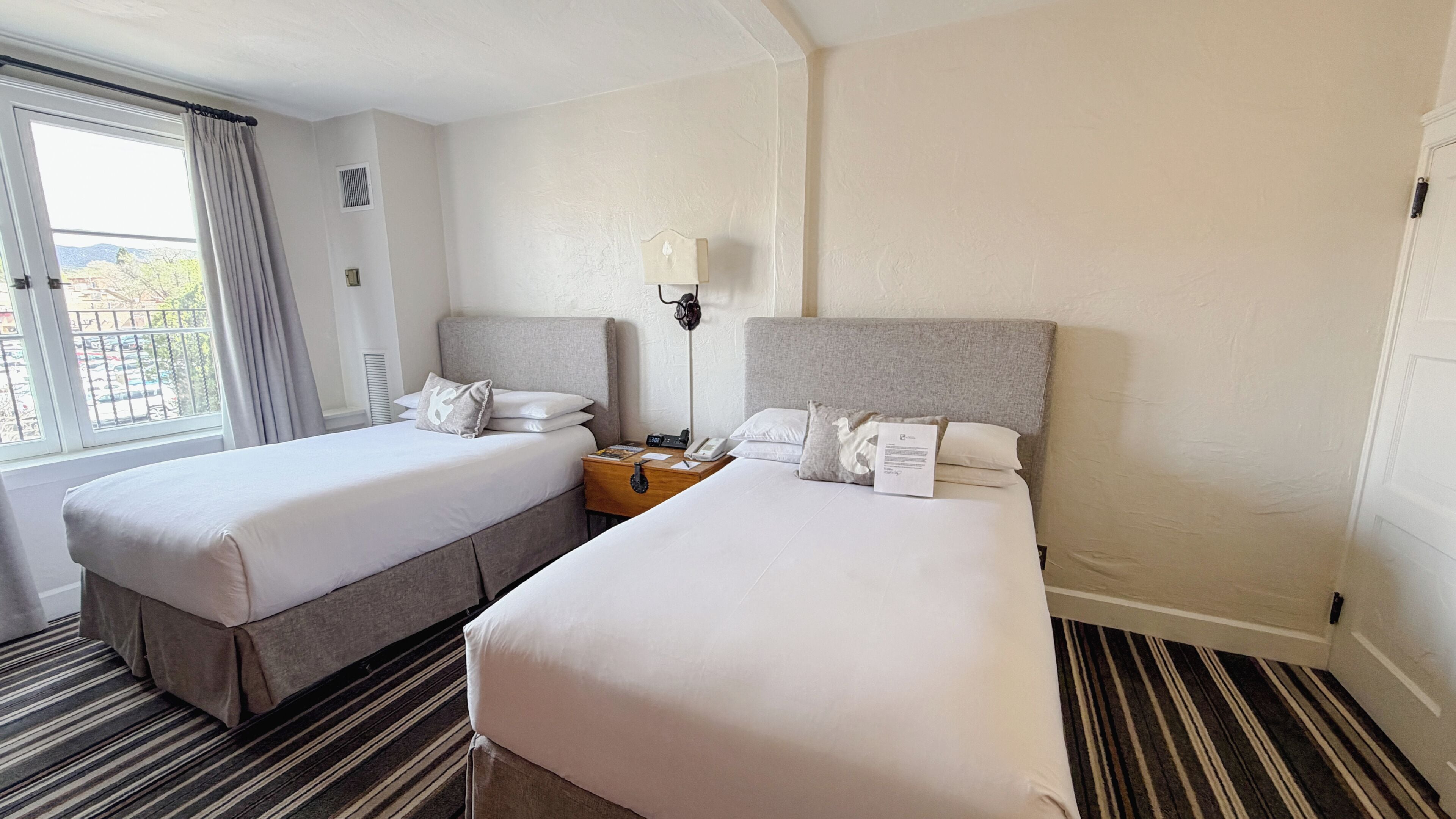 Une chambre d’hôtel avec deux lits, une table de chevet avec une lampe, une fenêtre avec des rideaux et un tapis à rayures.
