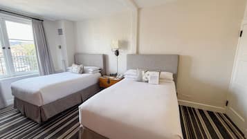 Habitación de hotel con dos camas, una mesita de noche con lámpara, una ventana con cortinas y una alfombra a rayas.