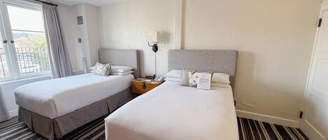 Une chambre d’hôtel avec deux lits, une table de chevet avec une lampe, une fenêtre avec des rideaux et un tapis à rayures.