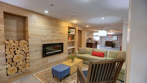 A living room with a TV, a sofa, a coffee table, and a chair. There is a stack of firewood on the left and a bookshelf with various items.