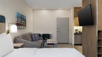 Camera d'albergo moderna con un letto grande, una poltrona, un tavolino, una scrivania, una televisione a schermo piatto e un'uscita che conduce a un angolo cottura.