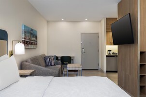 Una habitación de hotel moderna con una cama grande, un sofá, una mesita, un escritorio, un televisor de pantalla plana y una puerta que da a una kitchenette.