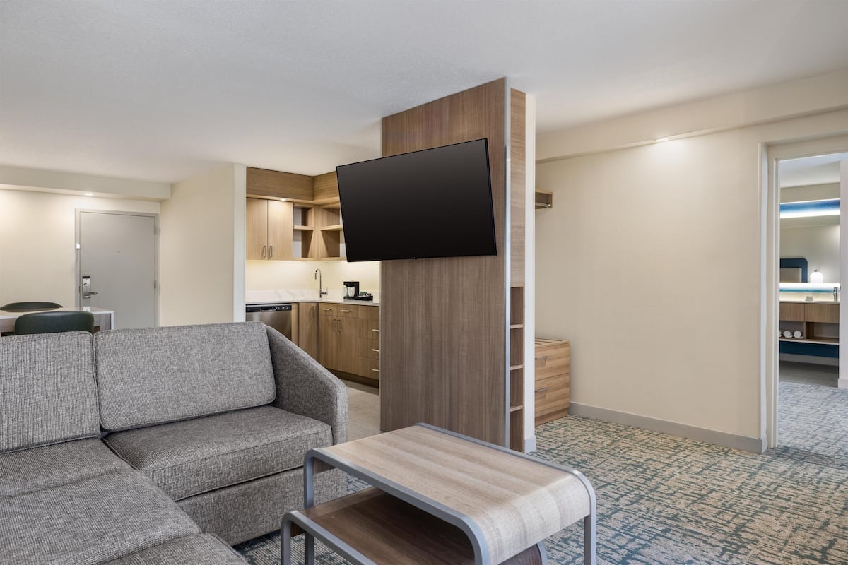 A modern hotel room with a sofa, a small table, a flat-screen TV mounted on a wooden panel, a kitchenette, and a bathroom visible through an open door.