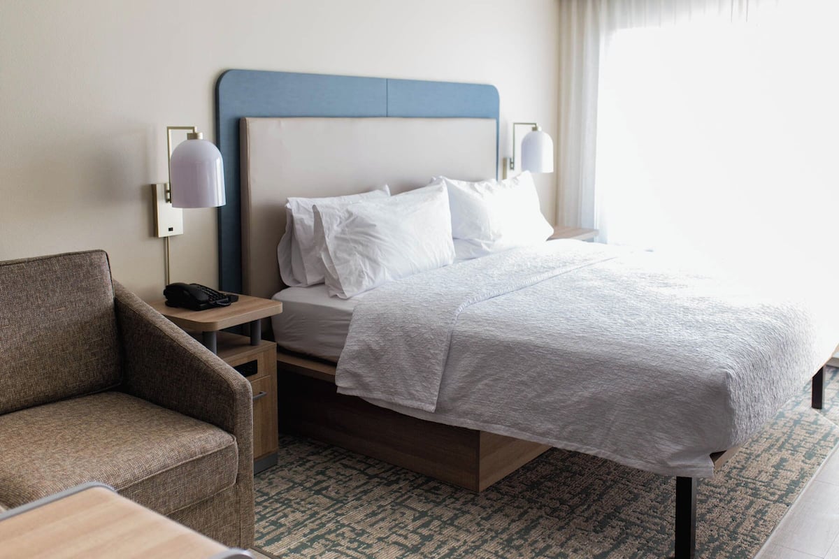 A hotel room with a bed, a chair, a bedside table with a lamp, and a wall-mounted phone.