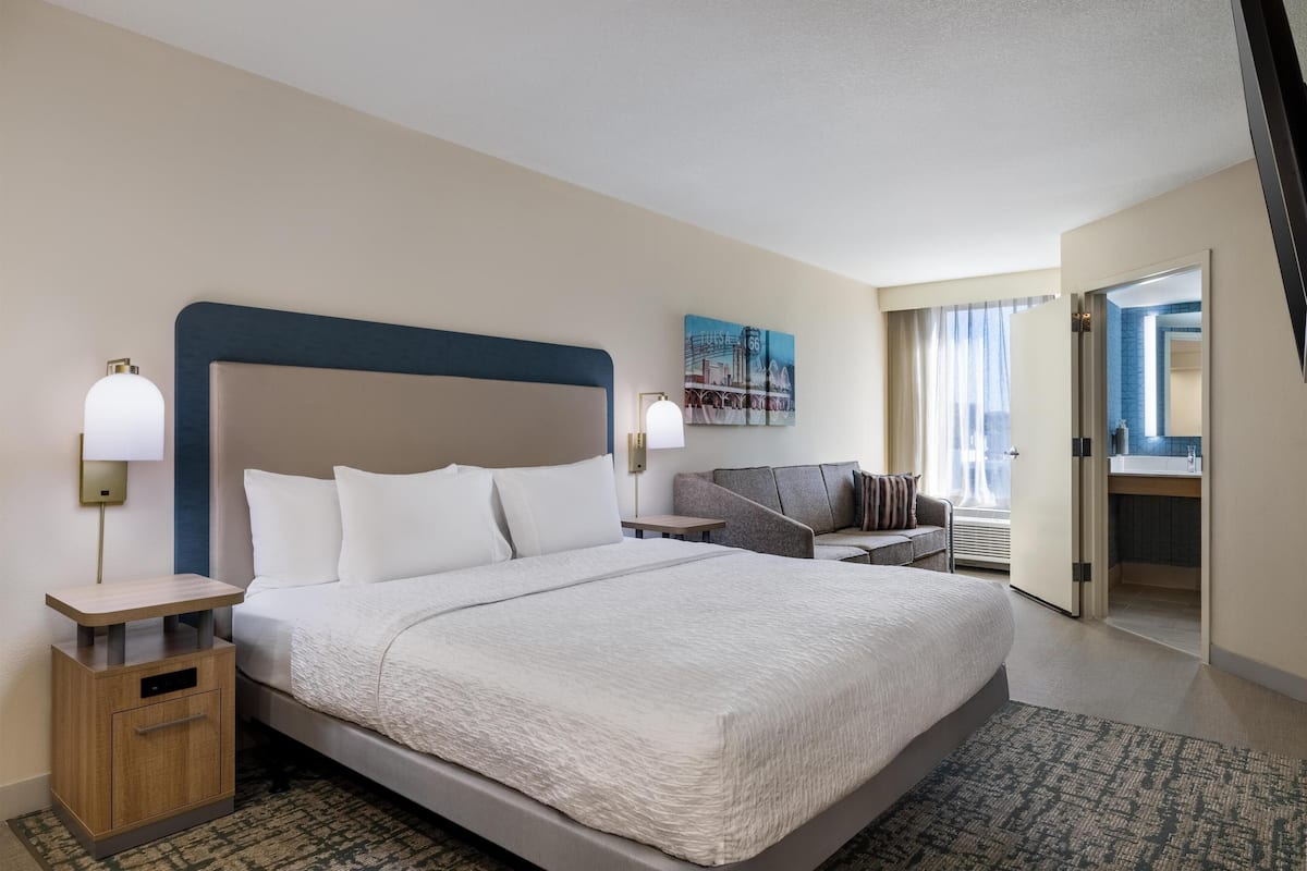 A hotel room with a large bed, a bedside table, a chair, a desk, and a bathroom visible through an open door.