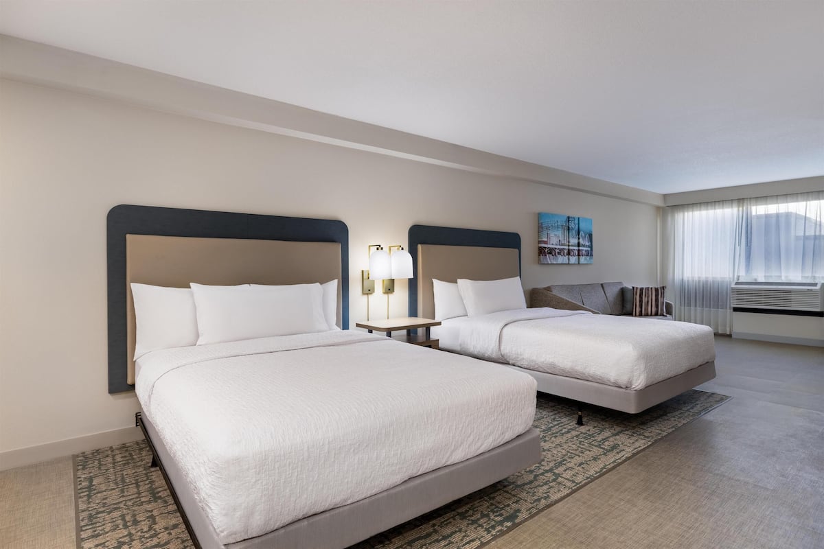 A hotel room with two beds, a nightstand, a lamp, a painting, and a large window with curtains.