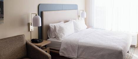 Ein Hotelzimmer mit Bett, Sessel, Nachttisch, Lampe und einem fensterbepflanzten Fenster.