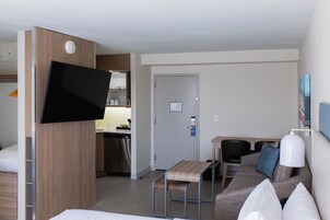 Habitación de hotel con cama, televisión montada en un panel de madera, kitchenette, una mesita, sofá y lámpara.