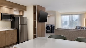 A modern kitchen with a microwave, refrigerator, and wooden cabinets. There is a flat-screen TV mounted on the wall, a dining area with a table and chairs, and a seating area with a sofa and a window with curtains. - Homewood Suites by Hilton Tulsa South Medical District (Tulsa)