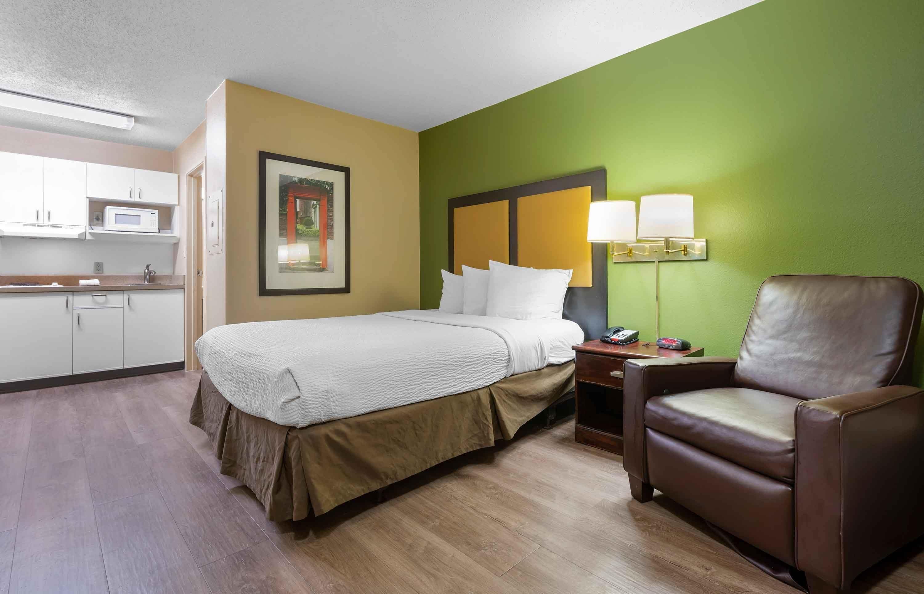 Une chambre d’hôtel équipée d’un lit, d’une chaise, d’une table de chevet, d’une lampe et d’un four à micro-ondes.