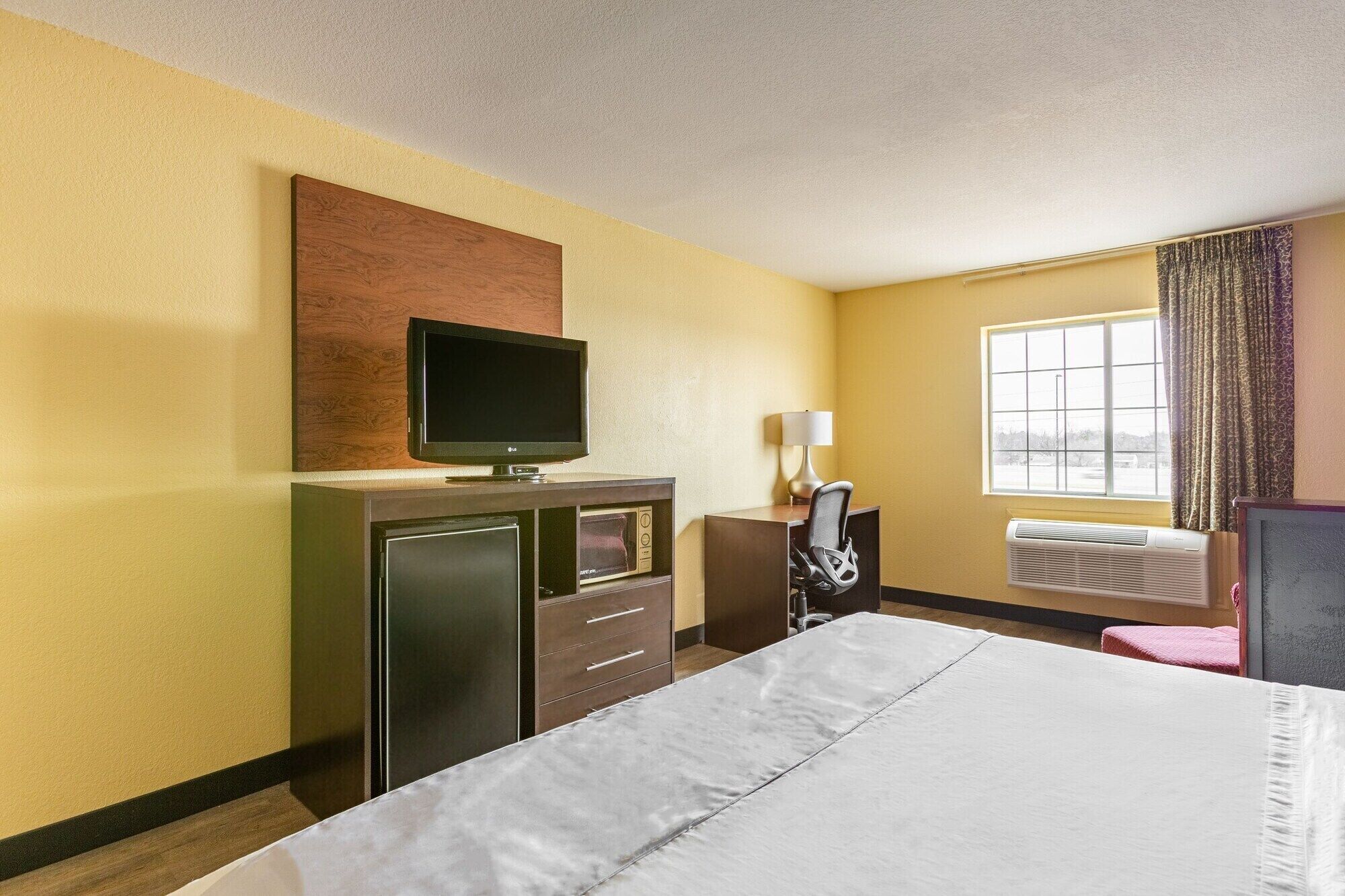 Habitación de hotel con cama, escritorio con silla, televisión, lámpara y ventana con cortinas.