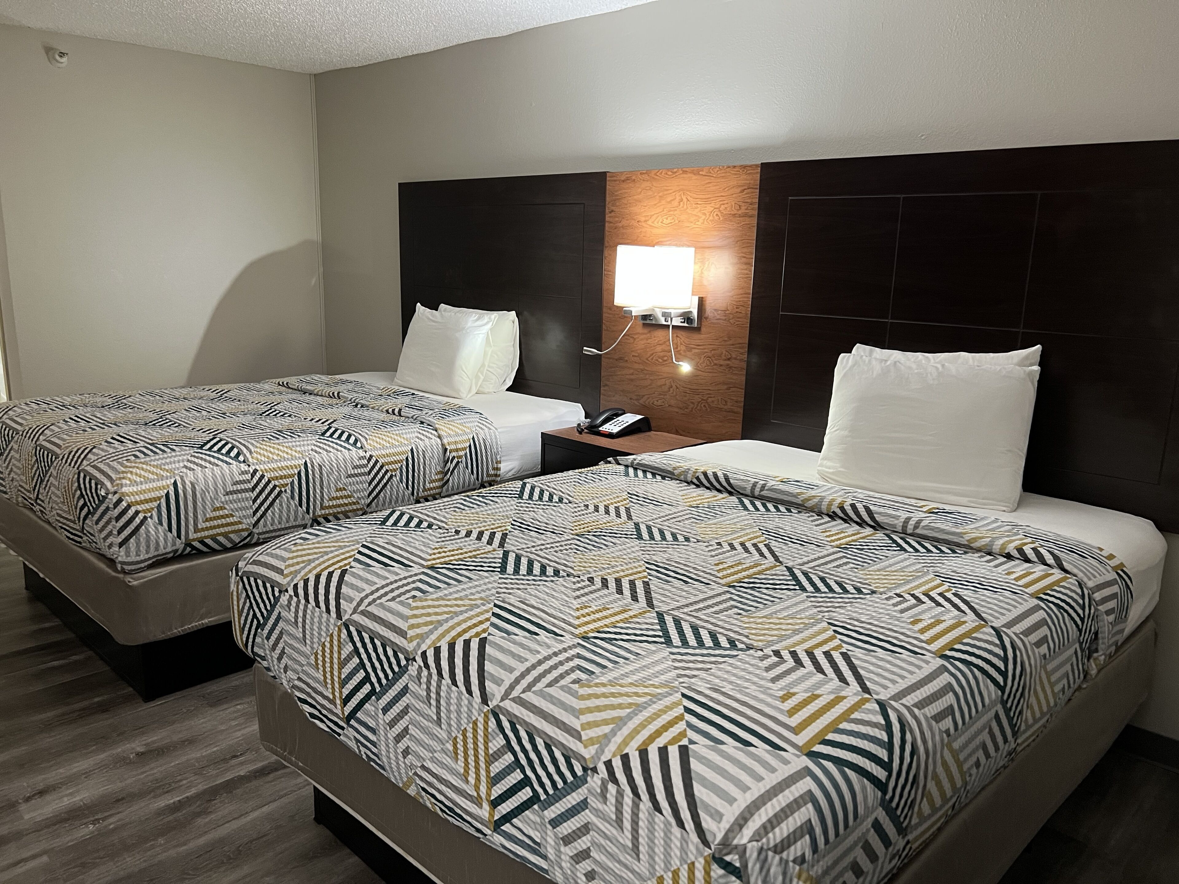 A hotel room with two beds, each with patterned bedding, a bedside table with a phone, and a wall-mounted light.