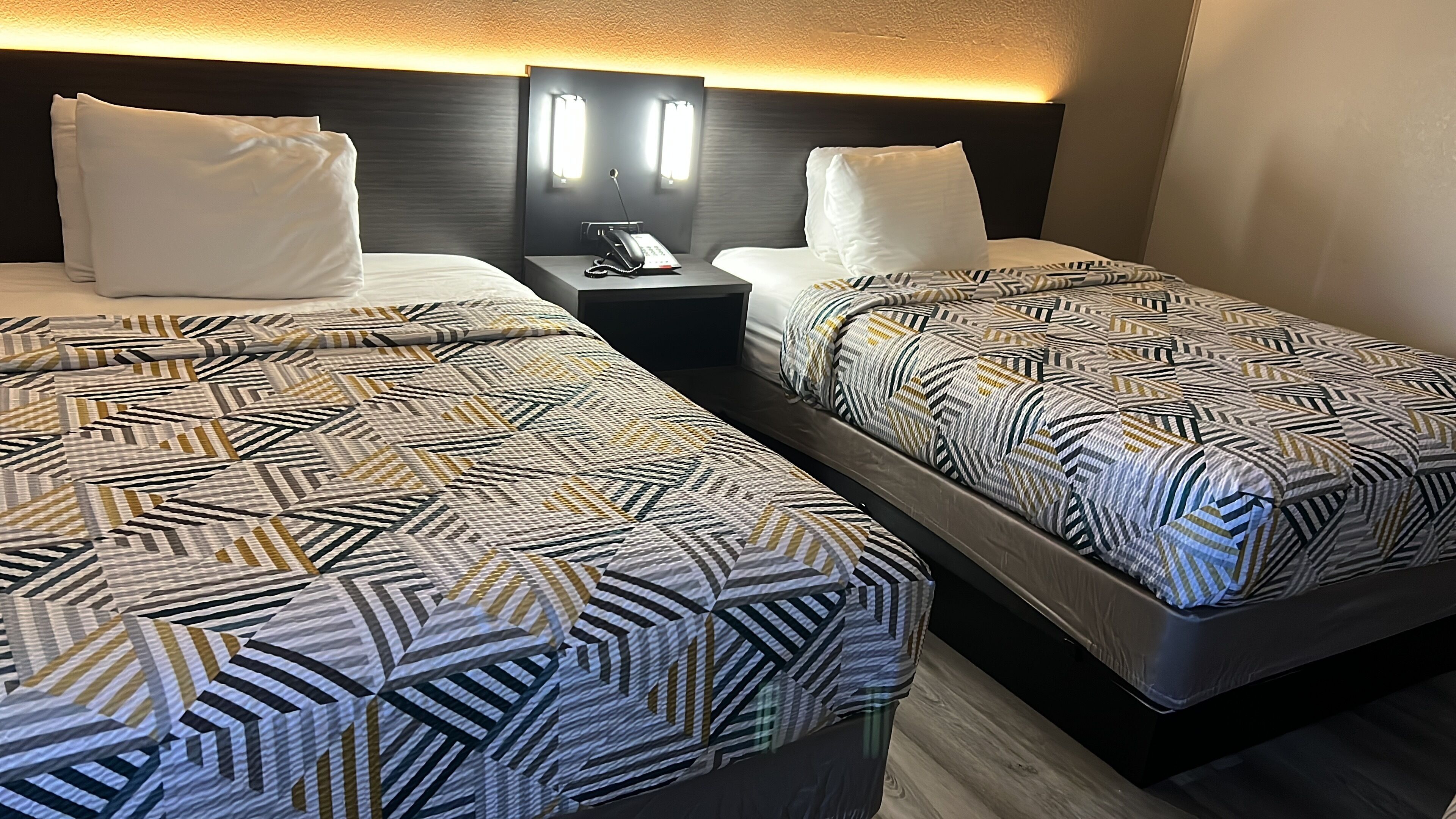 A hotel room with two beds, each with a patterned bedspread, a bedside table with a phone, and a wall-mounted light fixture.