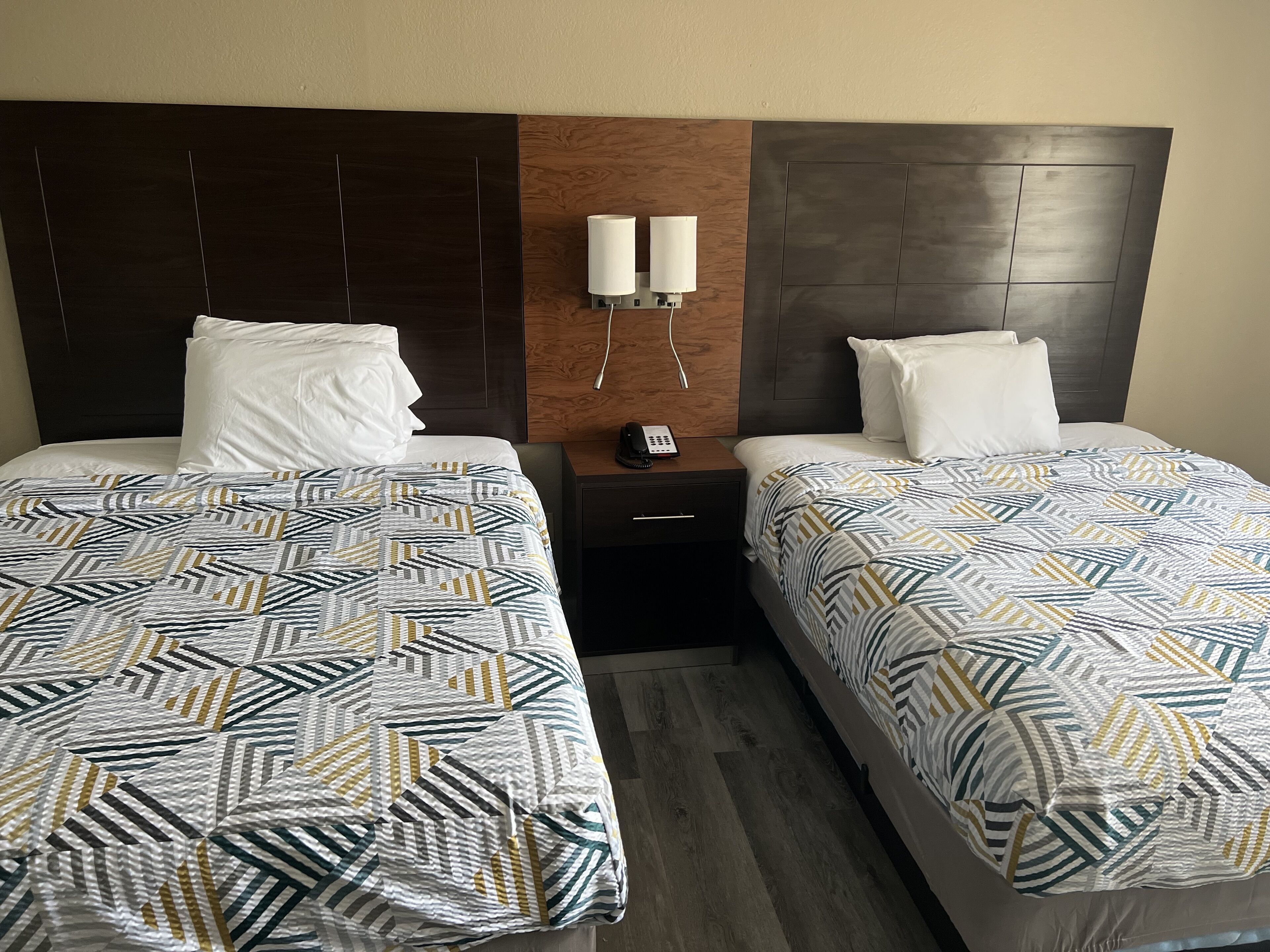 A hotel room with two beds, each with a patterned bedspread, a wooden headboard, a bedside table with a phone, and a wall-mounted light fixture.
