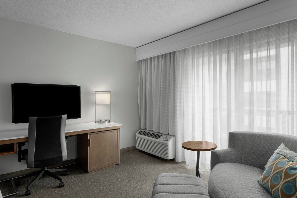 A hotel room with a flat-screen TV, a desk with a chair, a small table, a sofa, and a large window with sheer curtains.