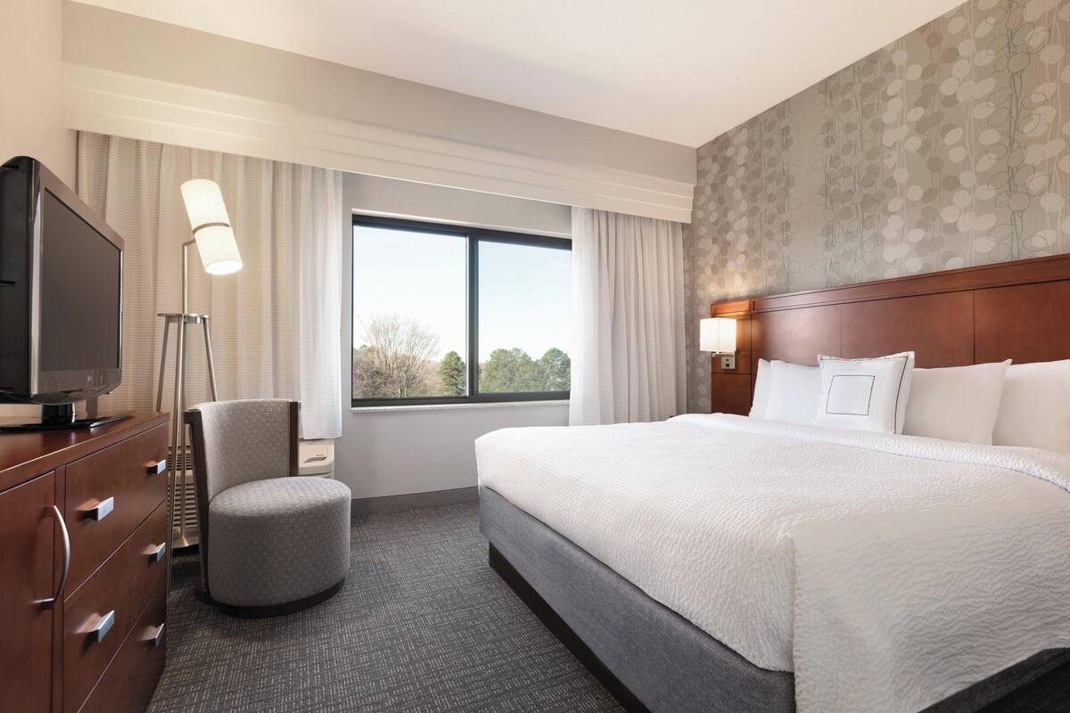 Habitación de hotel con una cama grande, un televisor en una cómoda, una silla y un ventanal con cortinas.