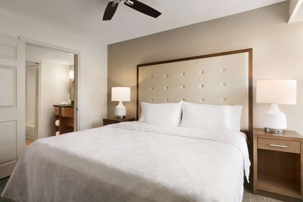 Ein Hotelzimmer mit einem großen Bett, zwei Nachttischen mit Lampen, einem Deckenventilator und einer Tür zu einem anderen Raum.