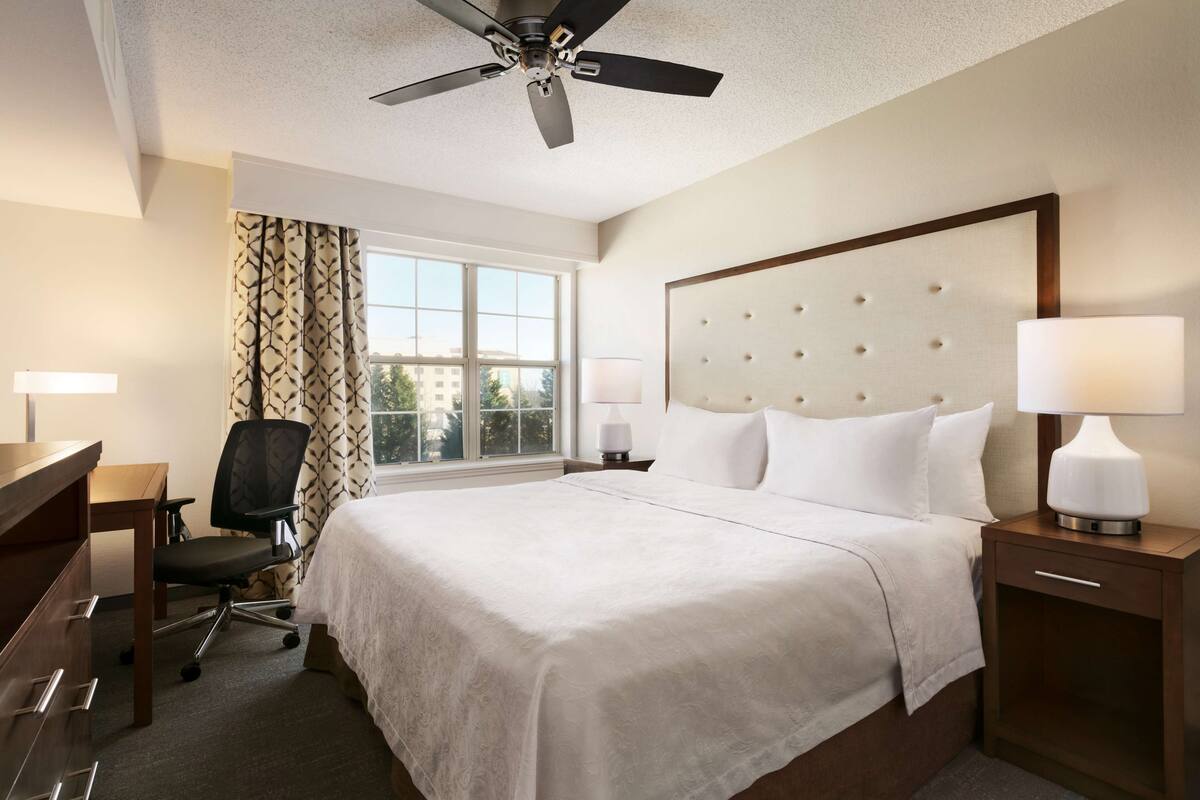 Ein Hotelzimmer mit einem großen Bett, einem Schreibtisch mit Stuhl, einem Deckenventilator und einem Fenster mit Vorhängen.