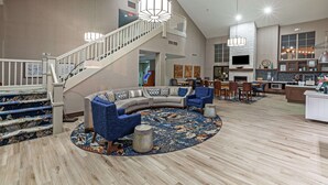 A spacious living area with a curved sofa, blue armchairs, a round coffee table, and a patterned rug. A staircase with a railing leads to an upper level.