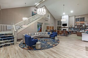 A spacious living area with a curved sofa, blue armchairs, a round coffee table, and a patterned rug. A staircase with a railing leads to an upper level.