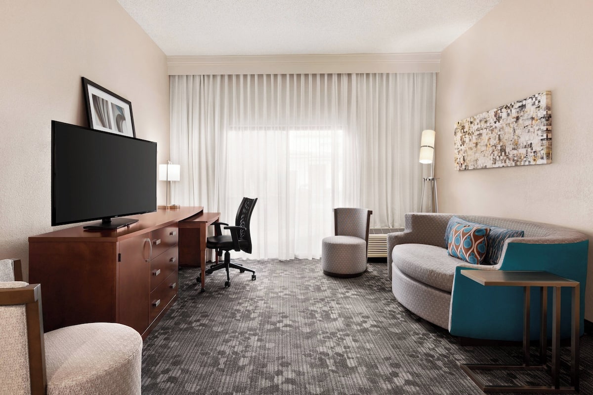 A hotel room with a flat-screen TV, a desk with a chair, a sofa with a side table, and a large window with curtains.