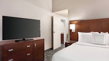 A hotel room with a large flat-screen TV on a wooden dresser, a neatly made bed with white bedding, and a wooden headboard.