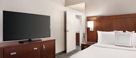 A hotel room with a large flat-screen TV on a wooden dresser, a neatly made bed with white bedding, and a wooden headboard.