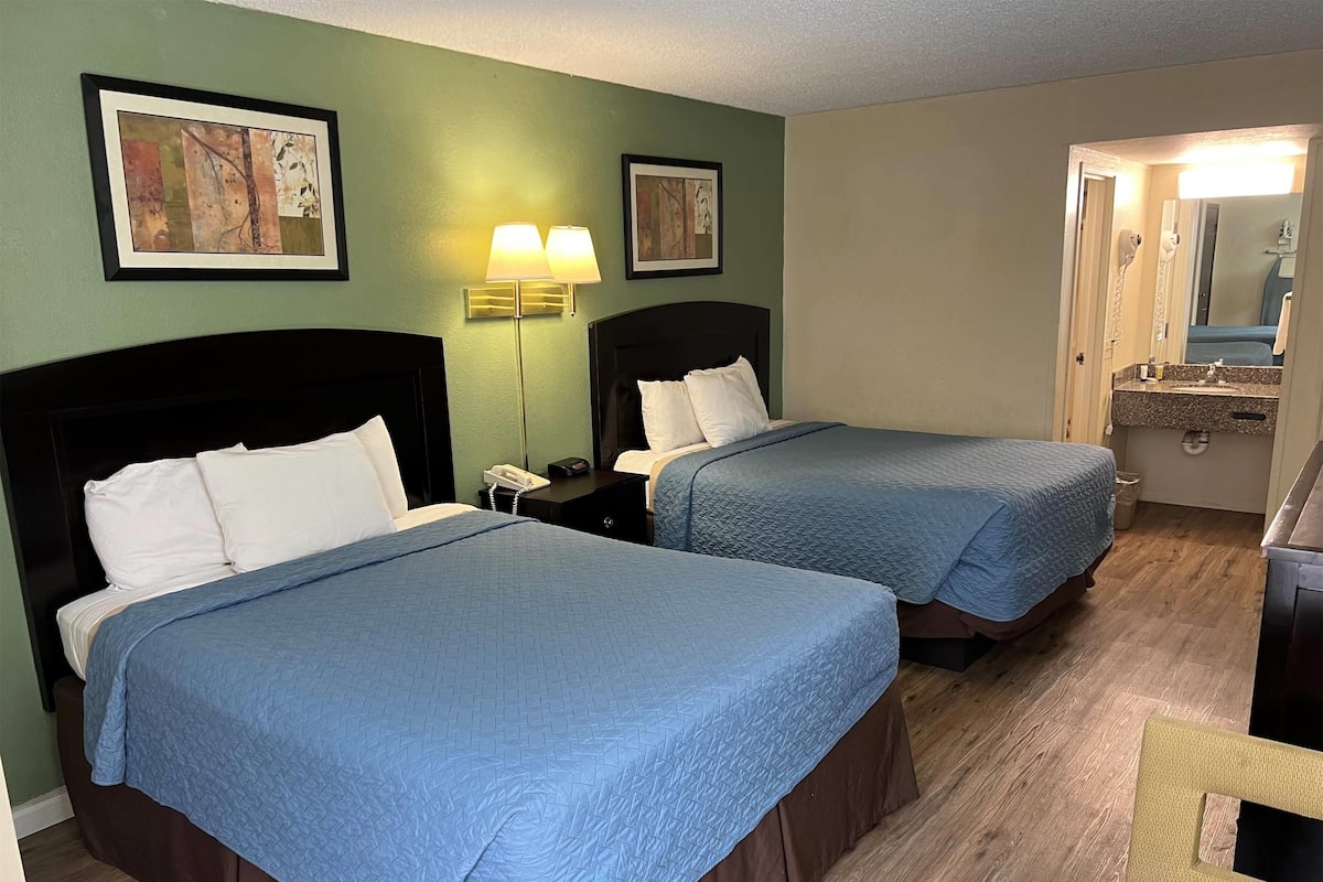 A hotel room with two beds, a nightstand with a lamp, and a bathroom visible through an open door.