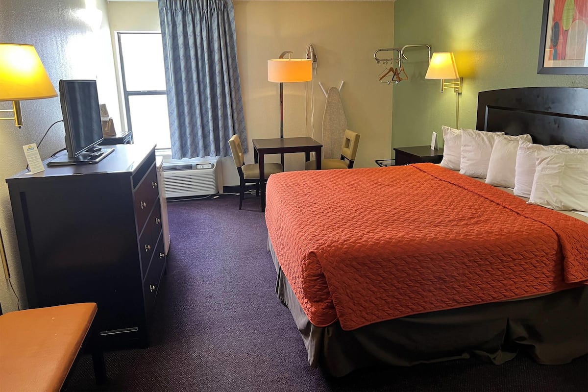 A hotel room with a bed, desk, chair, lamp, and air conditioning unit.