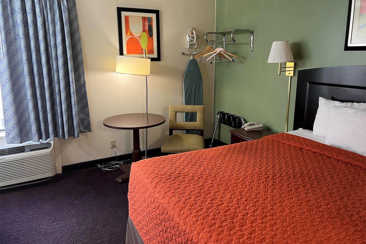 A hotel room with a bed, a chair, a small table, a lamp, and a wall-mounted coat rack.