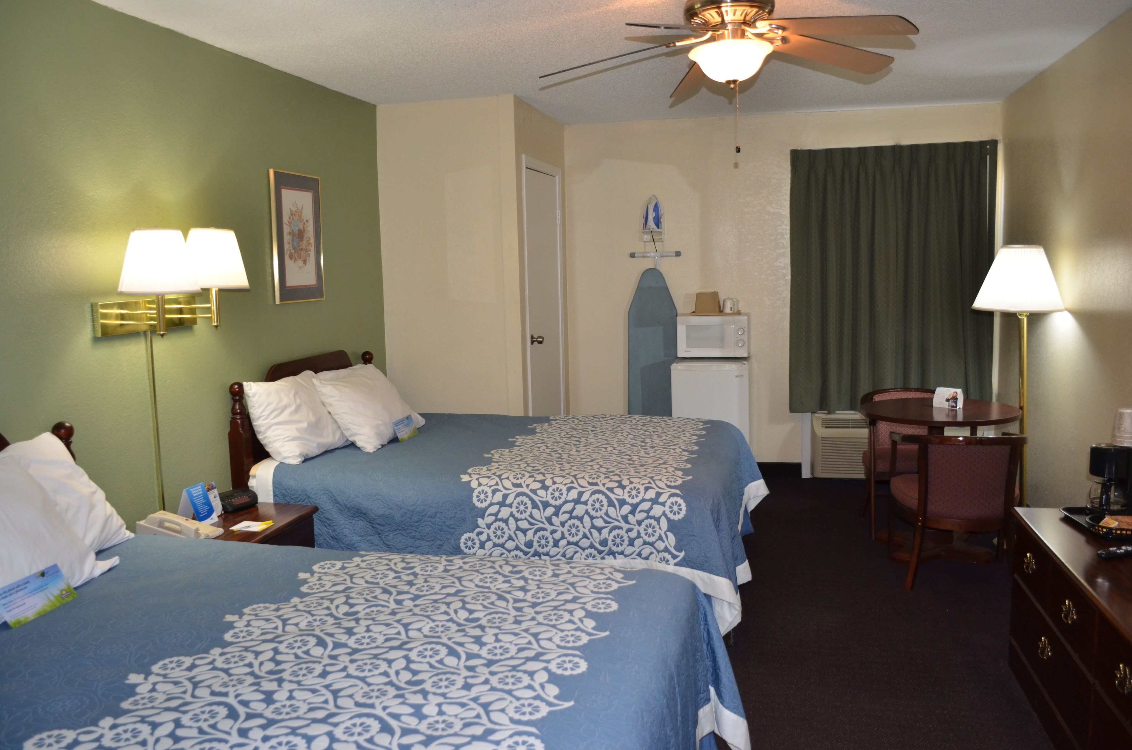 Une chambre d’hôtel avec deux lits, un bureau, une chaise, un ventilateur de plafond et une fenêtre avec des rideaux.