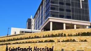 Ein modernes Krankenhausgebäude mit einem markanten Kreuz an der Fassade, einer Steinsäule mit Grünpflanzen und der Aufschrift 'SaintFrancis Hospital'.