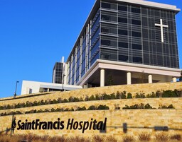 Un moderno edificio hospitalario con una cruz destacada en la fachada, un muro de piedra con vegetación y el nombre 'SaintFrancis Hospital' a la vista.