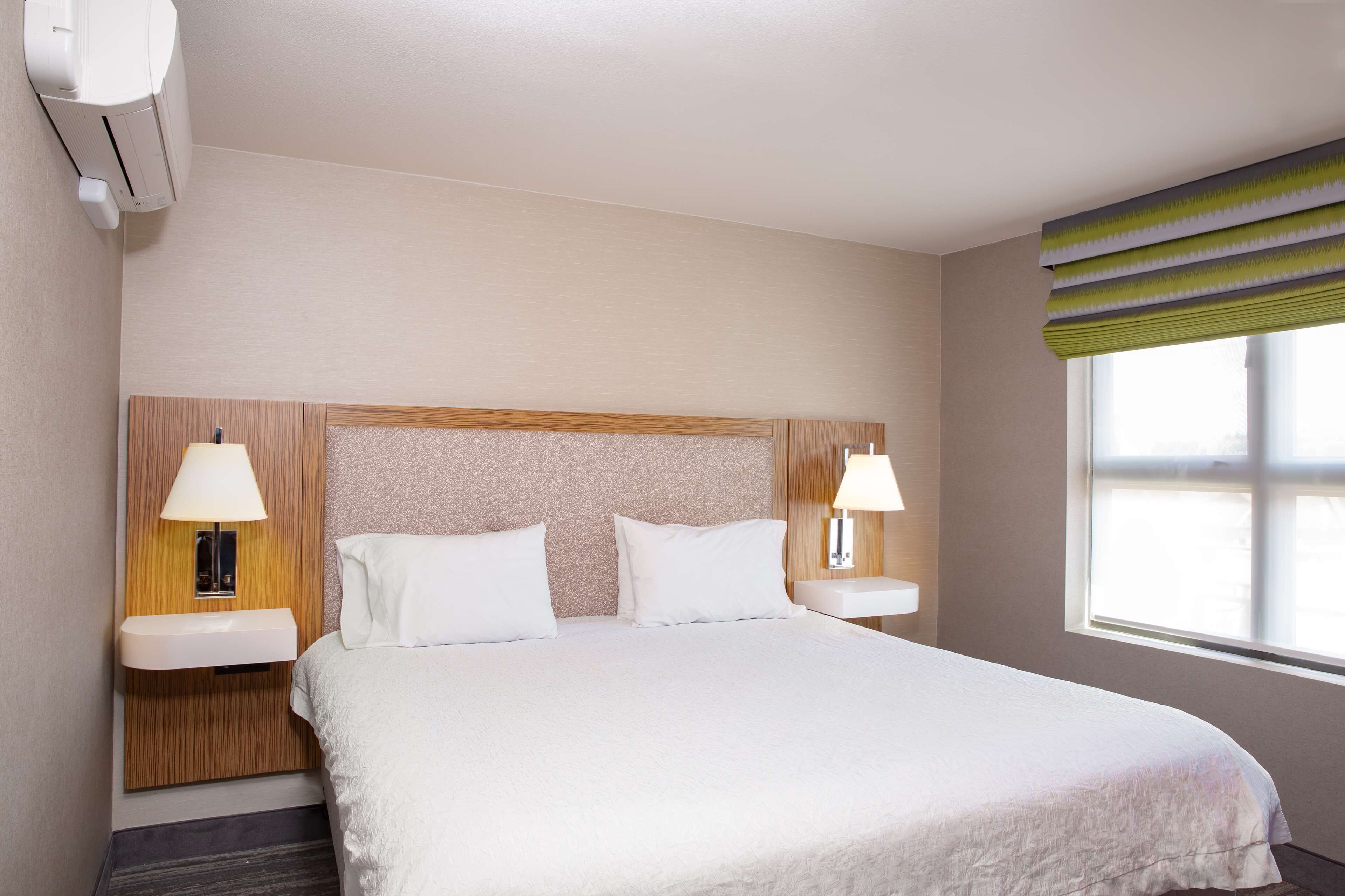 A hotel room with a large bed, two bedside tables with lamps, a wall-mounted air conditioner, and a window with a striped blind.