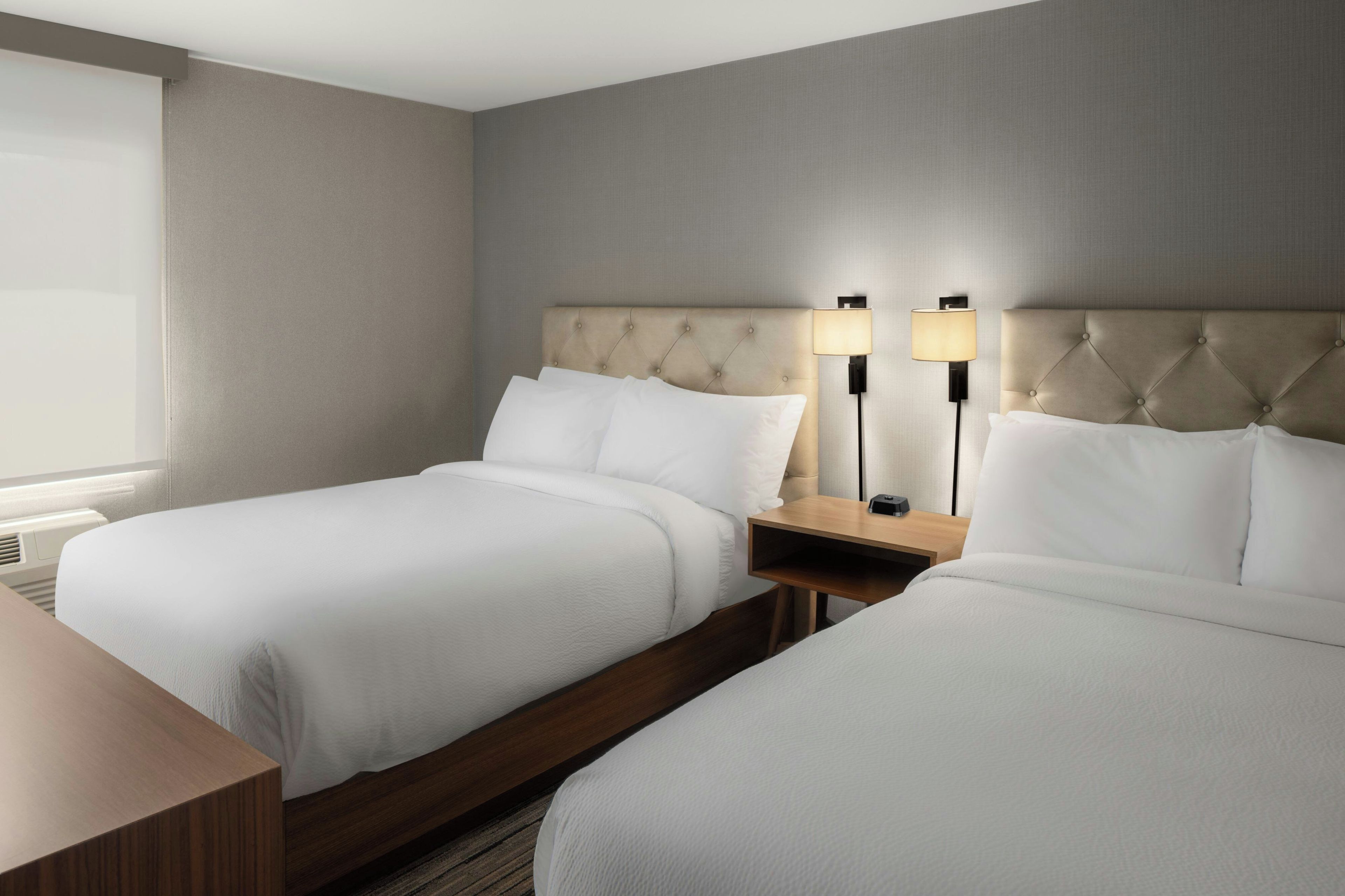 A hotel room with two beds, a nightstand, a lamp, and a window.