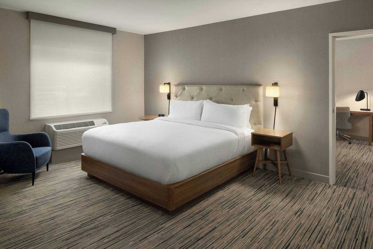 Una habitación de hotel con una cama grande, dos mesitas de noche con lámparas, una silla, una ventana con persianas y piso alfombrado.