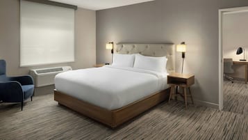 Habitació d'hotel amb un llit gran, dues taules de nit amb làmpades, una cadira, una finestra amb persianes i un sòl amb moqueta.