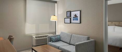 A hotel room with a grey sofa, a wooden coffee table, a bed, a wall-mounted air conditioner, and framed pictures on the wall.
