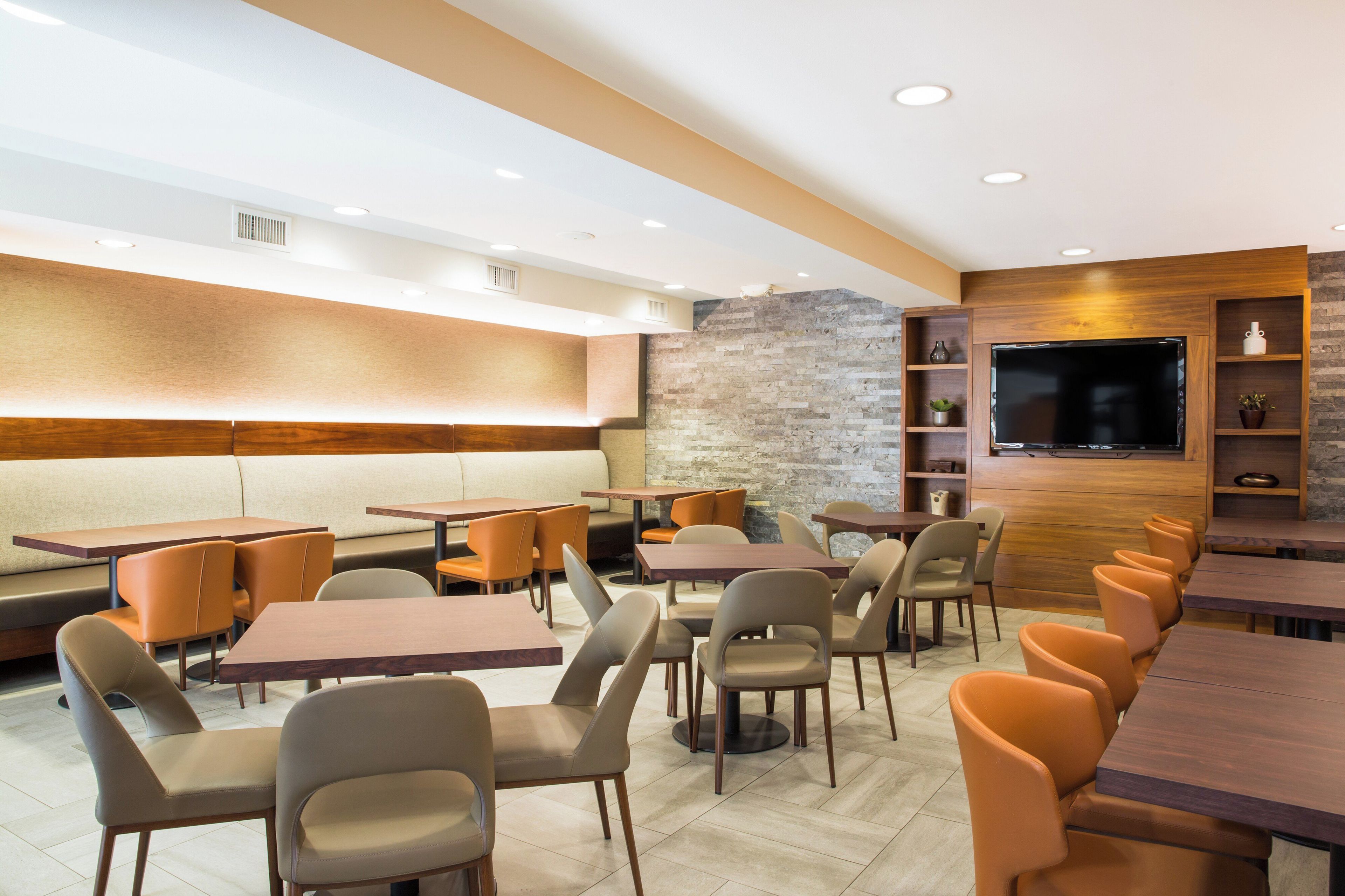 Interior moderno de restaurante com diversas opções de assento, incluindo boxes e mesas, parede com detalhes em pedra e uma TV de tela plana na parede.