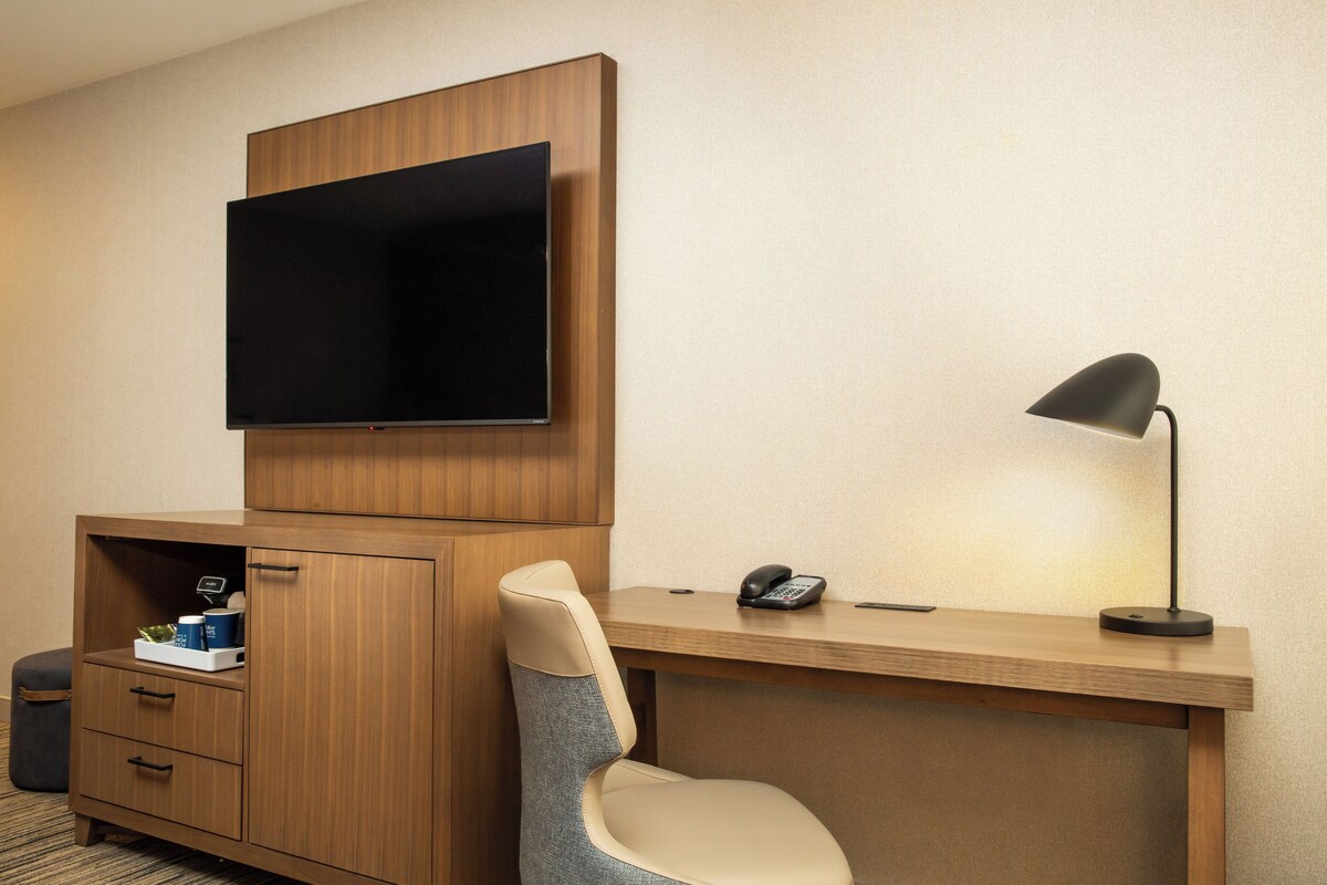 A hotel room with a wooden desk, a chair, a television mounted above the desk, a telephone, and a lamp.