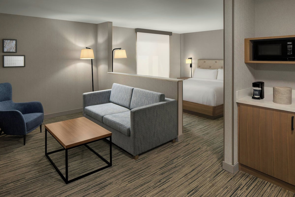 Habitación de hotel con un sofá gris, una mesa de centro de madera, un sillón azul, una cama con cabecero, una mesita de noche con lámpara, un microondas y un mueble pequeño.